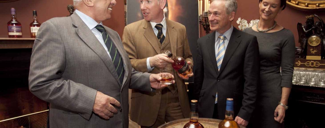 Vier Personen in formeller Kleidung neben einem Whiskeyfass in elegantem Raum.