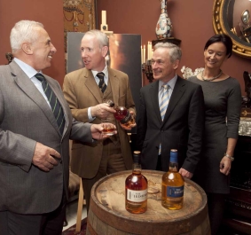 Vier Personen in formeller Kleidung neben einem Whiskeyfass in elegantem Raum.