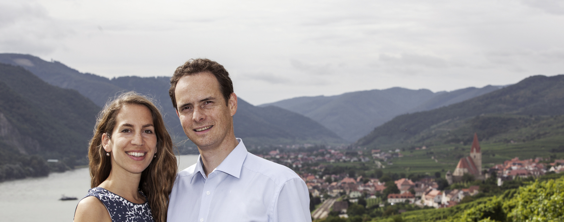Paar vor malerischer Berglandschaft mit Fluss und Weinbergen im Hintergrund.