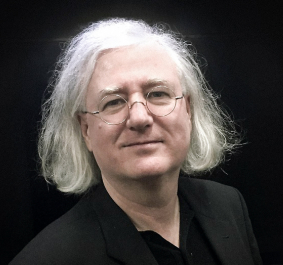 Älterer Mann mit weißem Haar und Brille vor schwarzem Hintergrund.