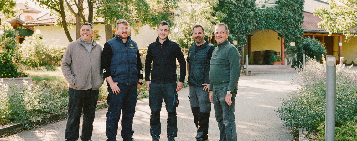 Fünf Männer lächeln vor einem idyllischen Gebäude im Grünen.