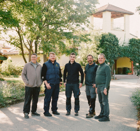 Fünf Männer lächeln vor einem idyllischen Gebäude im Grünen.