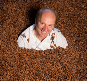 Mann in weißem Kittel, umgeben von vielen Kakaobohnen.