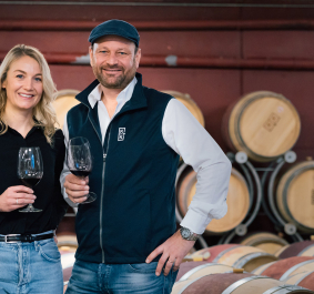 Zwei Personen mit Weingläsern in einem Weinkeller vor Holzfässern.