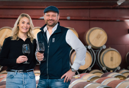 Zwei Personen mit Weingläsern in einem Weinkeller vor Holzfässern.