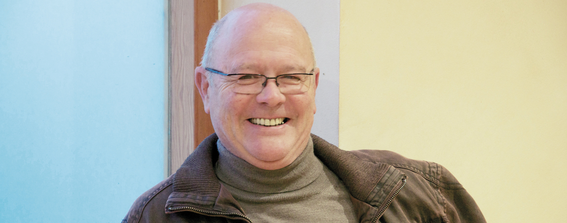 Lächelnde Person in brauner Jacke vor zweifarbiger Wand.