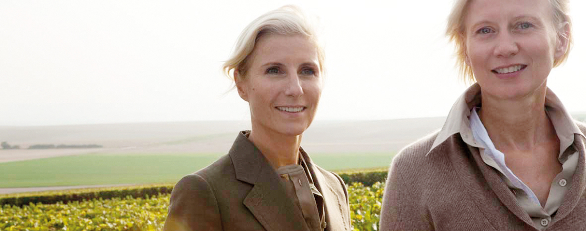 Zwei Frauen in eleganter Kleidung vor einer weitläufigen Landschaft mit Weinreben.