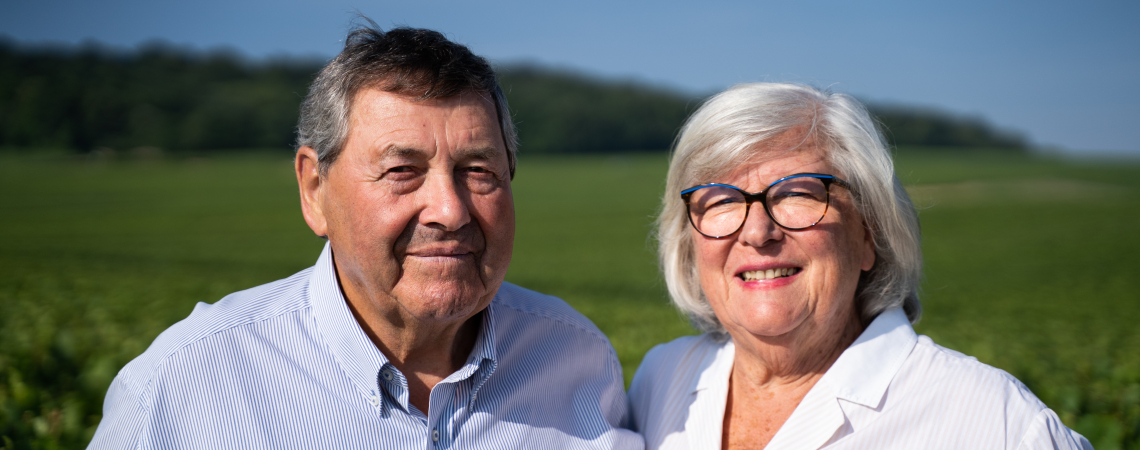 Älteres Paar mit weißen Hemden lächelt in grüner Landschaft.