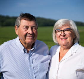 Älteres Paar mit weißen Hemden lächelt in grüner Landschaft.
