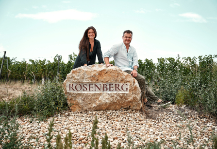 Zwei Personen sitzen lächelnd auf großem Felsen in Weinberg-Landschaft.