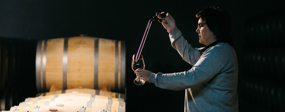 Person entnimmt Weinprobe aus Fass in dunklem Weinkeller.