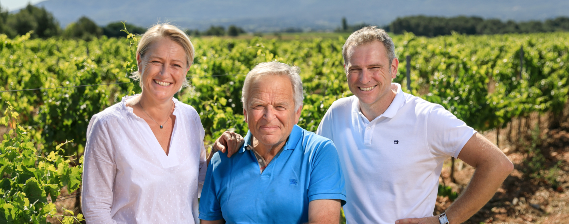 Drei lachende Personen im Weinberg mit Bergen im Hintergrund.