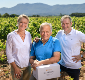 Drei lachende Personen im Weinberg mit Bergen im Hintergrund.