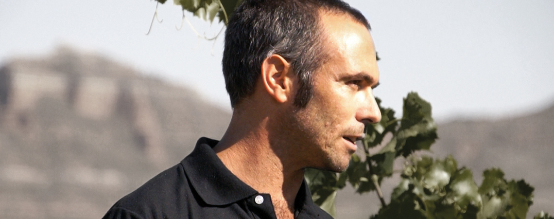 Mann im schwarzen Polohemd vor grünen Blättern und verschwommenem Bergpanorama.