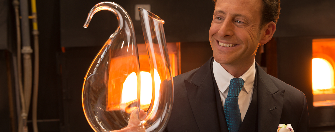 Mann in Anzug hält eleganten, geschwungenen Glasdekanter vor Ofenhintergrund.