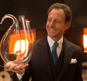 Mann in Anzug hält eleganten, geschwungenen Glasdekanter vor Ofenhintergrund.