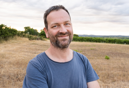 Mann im blauen T-Shirt steht lächelnd auf einem Feld vor Landschaftshintergrund.