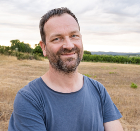 Mann im blauen T-Shirt steht lächelnd auf einem Feld vor Landschaftshintergrund.