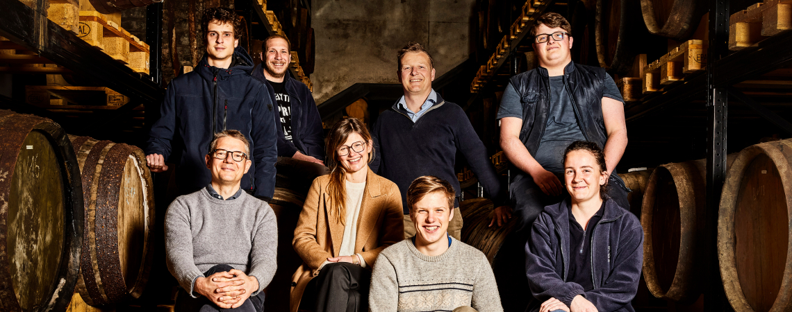 Gruppe von acht Menschen in Weinkeller mit Holzfässern im Hintergrund.