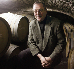 Mann in Weinkeller, umgeben von Holzfässern, trägt graues Sakko.