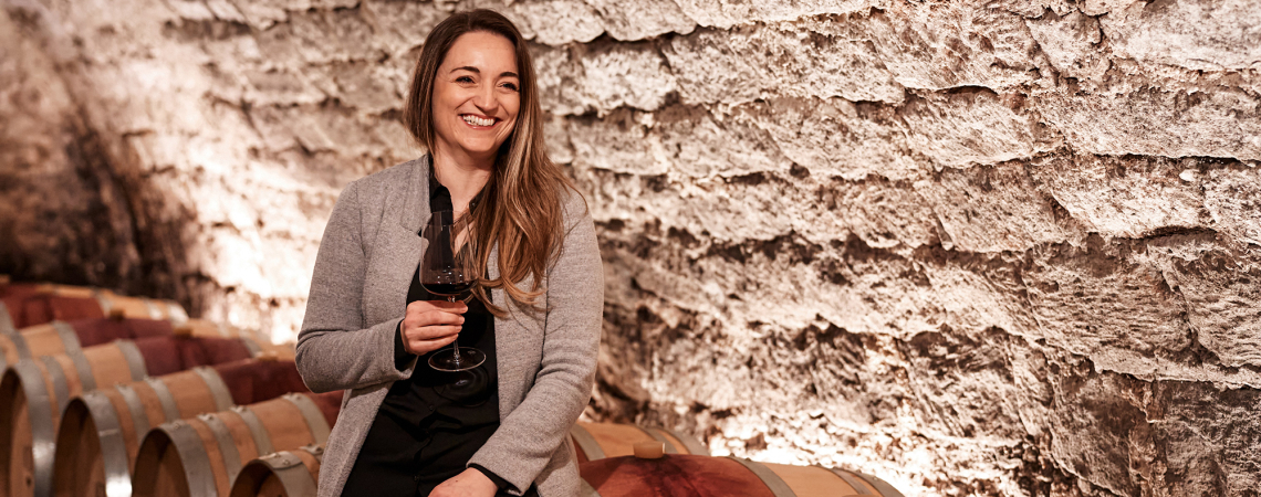 Frau in grauer Jacke sitzt lächelnd mit Weinglas auf Weinfass im Keller.