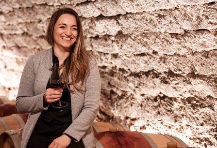 Frau in grauer Jacke sitzt lächelnd mit Weinglas auf Weinfass im Keller.