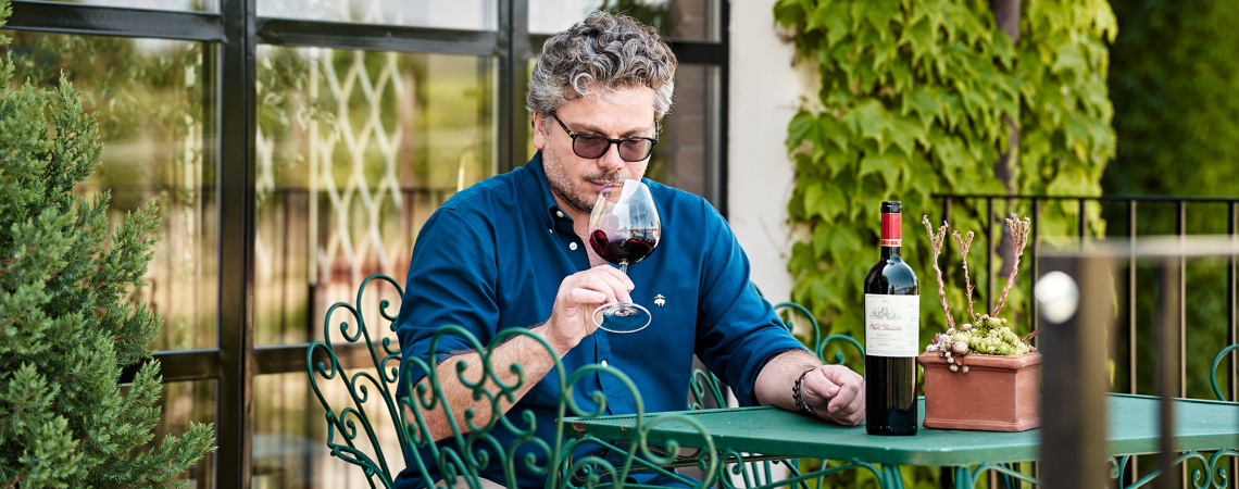 Mann in blauem Hemd genießt Rotwein auf Terrasse mit grünem Tisch.