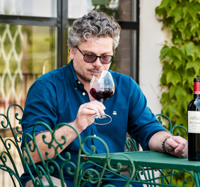 Mann in blauem Hemd genießt Rotwein auf Terrasse mit grünem Tisch.