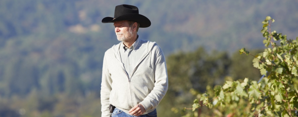 Mann mit Cowboyhut, grauem Pullover und Jeans spaziert durch Weinberge.