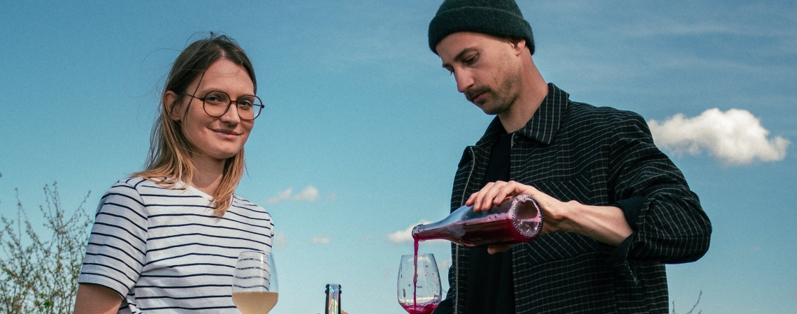 Zwei Personen mit Weingläsern und ein Hund auf einer Wiese unter blauem Himmel.