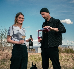 Zwei Personen mit Weingläsern und ein Hund auf einer Wiese unter blauem Himmel.