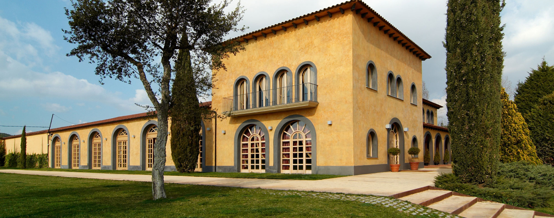 Gelbes Gebäude im mediterranen Stil mit Rundbogenfenstern und umgebenden Zypressen.