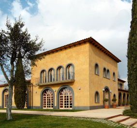 Gelbes Gebäude im mediterranen Stil mit Rundbogenfenstern und umgebenden Zypressen.