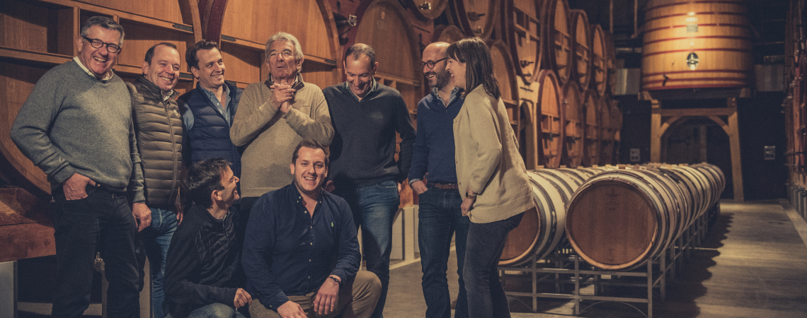 Gruppe von acht Menschen in einer Weinkellerei vor großen Holzfässern.