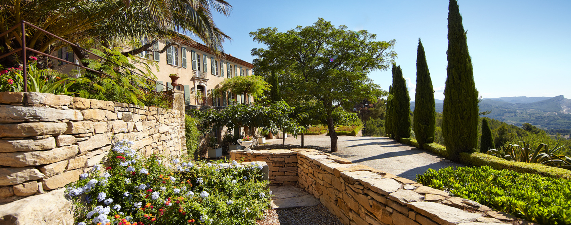 Mediterrane Villa mit Steinmauern, blühendem Garten und Zypressen im sonnigen Landschaftsbild.