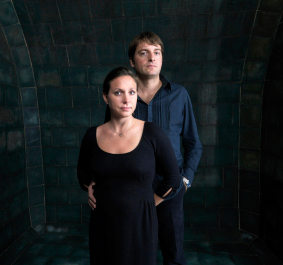 Zwei Personen in dunkler Kleidung vor einem dunklen, gekachelten Hintergrund.