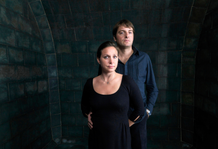 Zwei Personen in dunkler Kleidung vor einem dunklen, gekachelten Hintergrund.