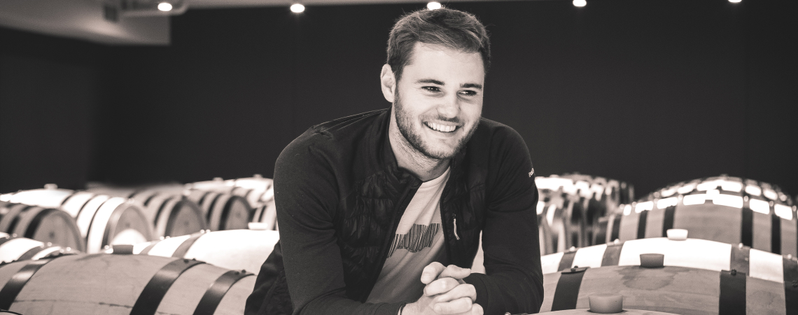 Mann lehnt sich lächelnd an Holzweinfässer in einem Kellerraum.
