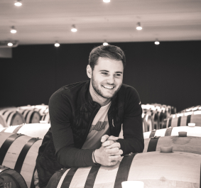 Mann lehnt sich lächelnd an Holzweinfässer in einem Kellerraum.