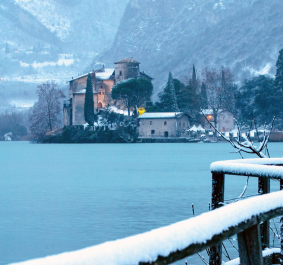 Verschneite Landschaft mit Schloss am Seeufer, gefrorene Äste und ein schneebedeckter Zaun im Vordergrund.