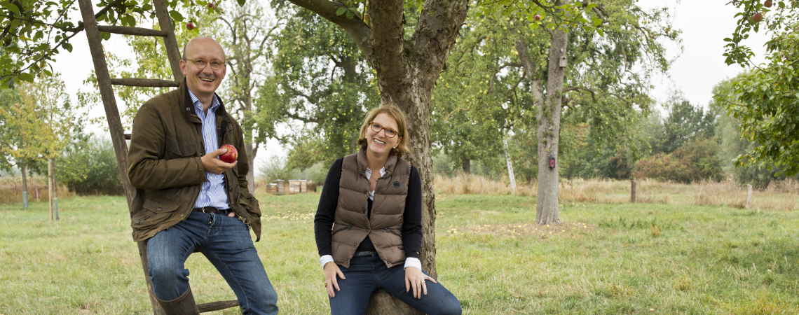 Zwei Personen auf einer Wiese, mit Körben voller Äpfel, vor einem Apfelbaum.