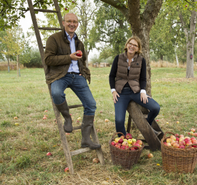 Zwei Personen auf einer Wiese, mit Körben voller Äpfel, vor einem Apfelbaum.