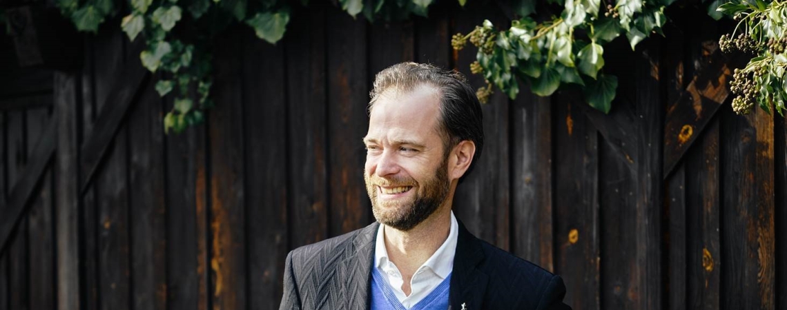 Mann in schwarzem Anzug und blauem Pullover vor Holzwand mit Efeu.