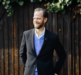 Mann in schwarzem Anzug und blauem Pullover vor Holzwand mit Efeu.