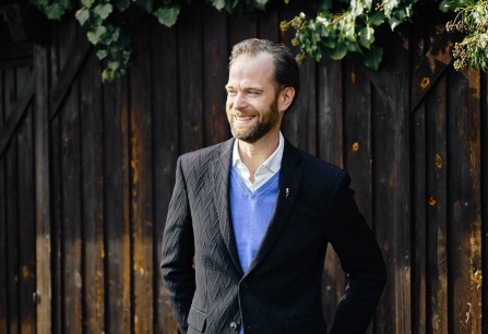 Mann in schwarzem Anzug und blauem Pullover vor Holzwand mit Efeu.