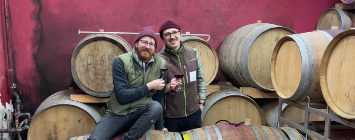 Zwei Männer mit Weingläsern stehen vor Fässern in einem Weinkeller.