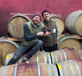 Zwei Männer mit Weingläsern stehen vor Fässern in einem Weinkeller.