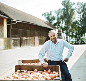 Mann in weißem Hemd sitzt auf Kiste mit Äpfeln vor Scheune.