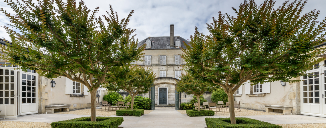 Elegante Steinvilla mit symmetrischem Garten und gepflastertem Innenhof, umgeben von Bäumen.
