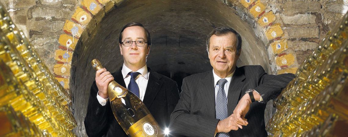 Zwei Männer in einem Weinkeller mit gestapelten Flaschen und großer Sektflasche.